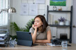 © Wasana - A woman is sitting at a desk with a laptop and a tablet. She is looking at the laptop and she is in a contemplative mood
