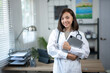© Wasana - A woman in a white lab coat is holding a laptop and smiling. She is in a medical setting, possibly a doctor's office or a hospital. Concept of professionalism and competence