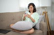 © Wasana - A woman is sitting on a couch with a pillow behind her back. She is using her cell phone and wearing headphones. Scene is relaxed and casual