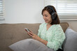 © Wasana - A woman is sitting on a couch with a cell phone in her hand. She is wearing headphones and she is listening to music