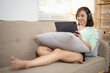 © Wasana - A woman is sitting on a couch with a tablet in her lap. She is wearing headphones and she is enjoying her time