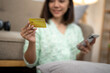 © Wasana - A woman is holding a credit card and looking at her phone. She is smiling and she is happy