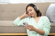 © Wasana - A woman is sitting on a couch with her phone in her hand. She is wearing headphones and she is listening to music