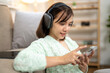 © Wasana - A woman is sitting on a couch and using her cell phone. She is wearing headphones and she is focused on her phone