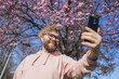 Happy curly man takes selfie against backdrop of flowering tree in spring for his internet communications. Weekend and social networks concept