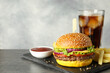 Burger with delicious patty, soda drink, french fries and sauce on dark table against gray background, space for text