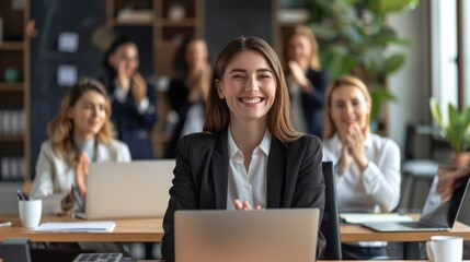 A smiling successful businesswoman sits at her desk with a laptop in her office, while a group of colleagues clap in the background.