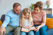 Happy multi-generational family reading a story book together sitting on sofa - Daughter, mother and grandfather having family time in the living room - Childhood concept