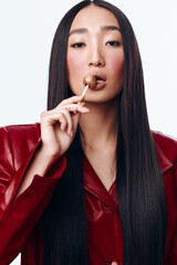 Wall Mural - Young Asian Woman with Long Black Hair and Red Jacket Enjoying a Lollipop in Studio Portrait Shot