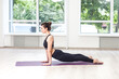 Full length portrait of slim woman working out doing yoga on firness mat, making cobra pose, relaxing, wearing black pants and top. Indoor shot in gym near big windows.