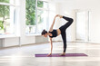 Full length portrait of athletic slim woman enjoying yoga, standing on one leg stretching her body, doing gymnastic exercising, wearing black pants and top. Indoor shot in gym near big windows.