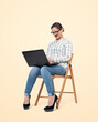 A young girl in glasses, shirt and jeans is sitting on a chair and working on a laptop, isolated on a bright yellow background