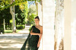 Young, beautiful woman in elegant clothes leans on a column in a park in Seville. In the background large trees on a sunny day. Holiday and travel concept.
