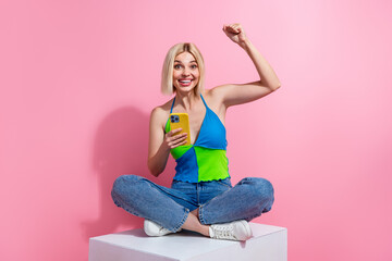 Wall Mural - Full length photo of ecstatic girl dressed colorful top denim pants sit on cube hold smartphone win bet isolated on pink color background