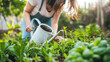 A woman is using a watering can to water plants in a lush garden setting.