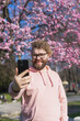 Spring day. Bearded man in pink shirt talking by phone. Spring pink sakura blossom. Handsome young man with smartphone. Fashionable man in trendy glasses. Bearded stylish man. Copy space