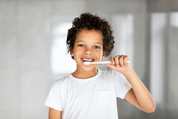 African American young boy with short hair is diligently brushing his teeth with a blue toothbrush in front of a mirror. He is standing in a bathroom, focused on his oral hygiene routine.