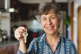 Fototapeta  - Joyful senior woman with short gray hair presents a key while standing in her home, symbolizing ownership, real estate investment, or new beginnings for the elderly