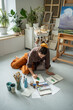 Concentrated woman artist painting with watercolor. Creative girl sitting on floor of home studio art workshop wielding brush creating sketches artworks with aquarelle paints working on draft project