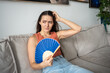 Disgruntled woman suffers from stuffiness, blowing by fan. Girl sitting on couch, feels discomfort from unbearable abnormal heat at home, scratches head. Woman sweating, dying from hot weather.