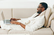 Young African American man working on a laptop in his cozy modern apartment He is sitting on the sofa, wearing a bathrobe, and typing away with a smile on his face It's a bright morning, and he is