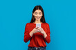A woman wearing a red shirt is holding a cell phone in her hand, looking at the screen. She appears focused on whatever is displayed on the device, possibly texting or browsing online.