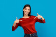 A smiling young woman, wearing a casual red long-sleeved top, stands in front of a solid blue backdrop. With a look of self-assuredness, she points both thumbs toward herself
