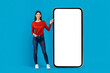 A smiling young woman dressed in a red top and blue jeans stands confidently next to an oversized smartphone model. She gestures towards the blank screen of the phone, mockup, copy space
