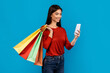A woman standing holding shopping bags in one hand and a cell phone in the other hand. She appears to be multitasking, possibly checking her phone while shopping, blue background