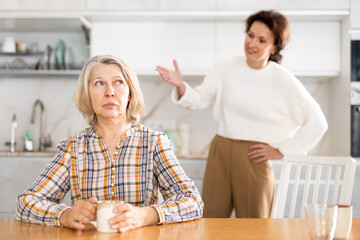 Wall Mural - Offended senior woman sitting in the kitchen holding jar while middle-aged woman talking angrily to her