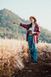 © Satori Studio - Smiling woman standing in a wheat field, holding a tablet, and pointing to the side, wearing a straw hat.
