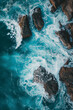 A breathtaking aerial view captures the vibrant turquoise ocean waves crashing against the jagged, weathered rocks, depicting the raw power and beauty of nature in this stunning seascape.