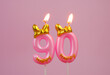 Burning pink birthday candle with gold bow and letters happy on pink background. Number 90.	