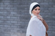 Portrait of a young blue-eyed woman in a hijab against a gray brick wall. A Muslim woman looks at the camera turning around.