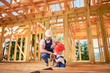 Father with toddler son building wooden frame house. Builder teaching his son how to use screwdriver on construction site, wearing helmets and blue overalls on sunny day. Carpentry and family concept.