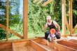 Father with toddler son building wooden frame house. Man playing with his son using electric saw, demonstrating that it unplugged from power source on construction site. Carpentry and family concept.