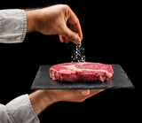 Fototapeta  - Chef is salting or seasoning raw ribeye steak laying on graphite serving board. Black background.