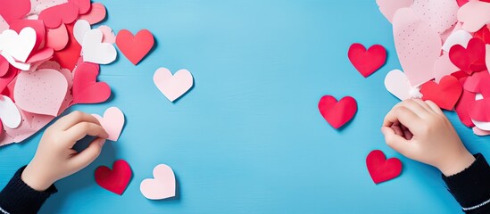Wall Mural - A top view of a child s hands cutting paper near various sized red and pink hearts on a blue background with plenty of empty space for text or other images. Copy space image