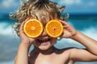 Happy child boy on the beach, laughing covering his eyes with orange halves, summer vacation concept