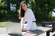 © MM - A woman in a white shirt and white pants sits on a park bench with a laptop in her hands.