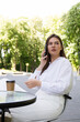 © MM - A woman in a white shirt and white pants sits at a table in a street cafe and talks on the phone.