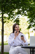 © MM - A woman in a white shirt and white pants sits at a table in a street cafe and talks on the phone.
