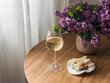 Home aperitif - a glass of white wine, cheese, olives, a pear and a bouquet of lilacs on a round wooden table in the living room