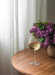 A glass of white wine and a bouquet of lilacs in a ceramic vase on a round wooden table