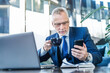 Senior male executive drinking coffee and holding smartphone while working online in office lounge. Business manager ceo using cell phone mobile app, laptop, computer
