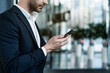 © InsideCreativeHouse - Cropped image of young businessman looking reading text message on his mobile phone standing in office lobby. Manager holding smartphone using mobile app. Digital, technology, business concept