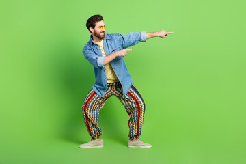 Sticker - Full length photo of cheerful handsome guy wear denim jacket dancing pointing empty space isolated green color background