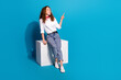 Photo portrait of nice young lady sit cube look point empty space wear trendy white garment isolated on blue color background