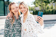 Two young beautiful smiling blond hipster female in trendy summer clothes. Sexy carefree women posing on street background. Positive models having fun, hugging at sunset. Cheerful and happy. Selfie