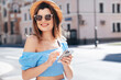 Young beautiful smiling woman in trendy summer clothes. Carefree female posing in street at sunny day. Positive model holds smartphone, looks at cellphone screen, uses mobile apps, in hat